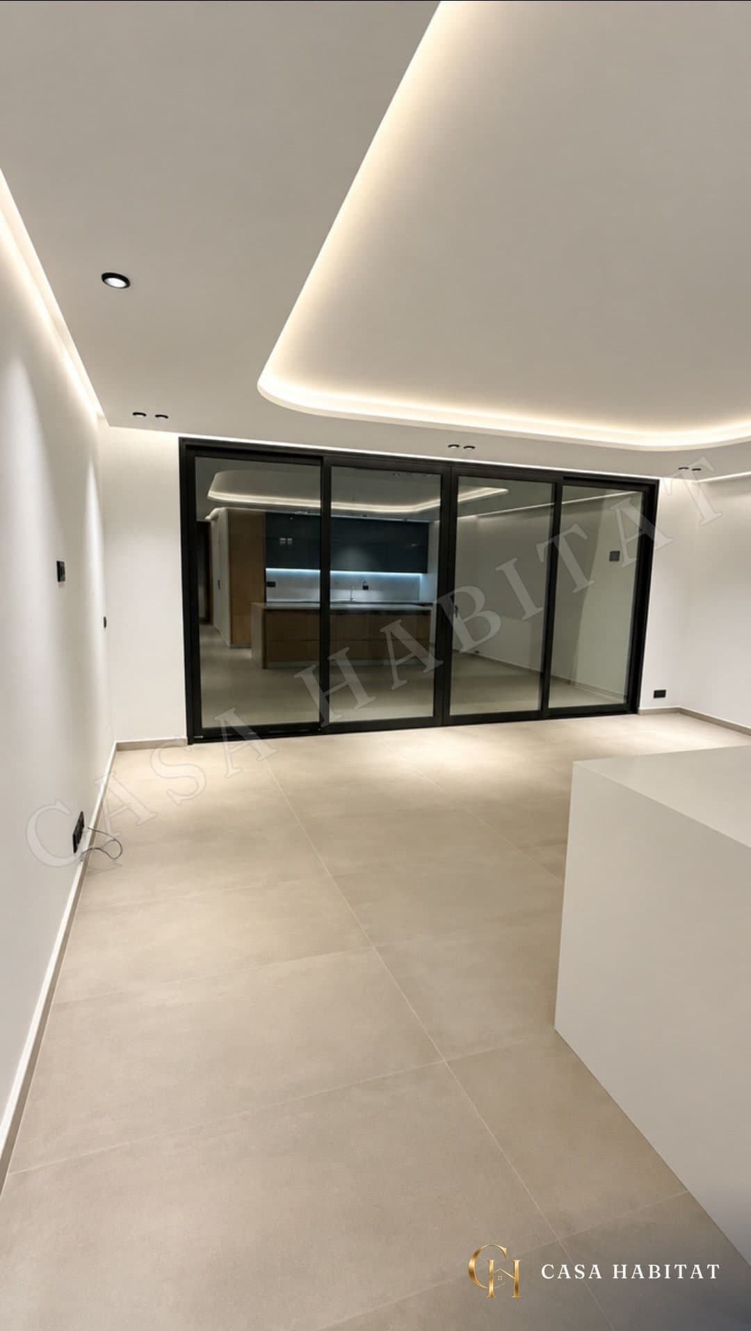 Salon vide au sol carrelé beige, grande baie vitrée donnant sur la cuisine