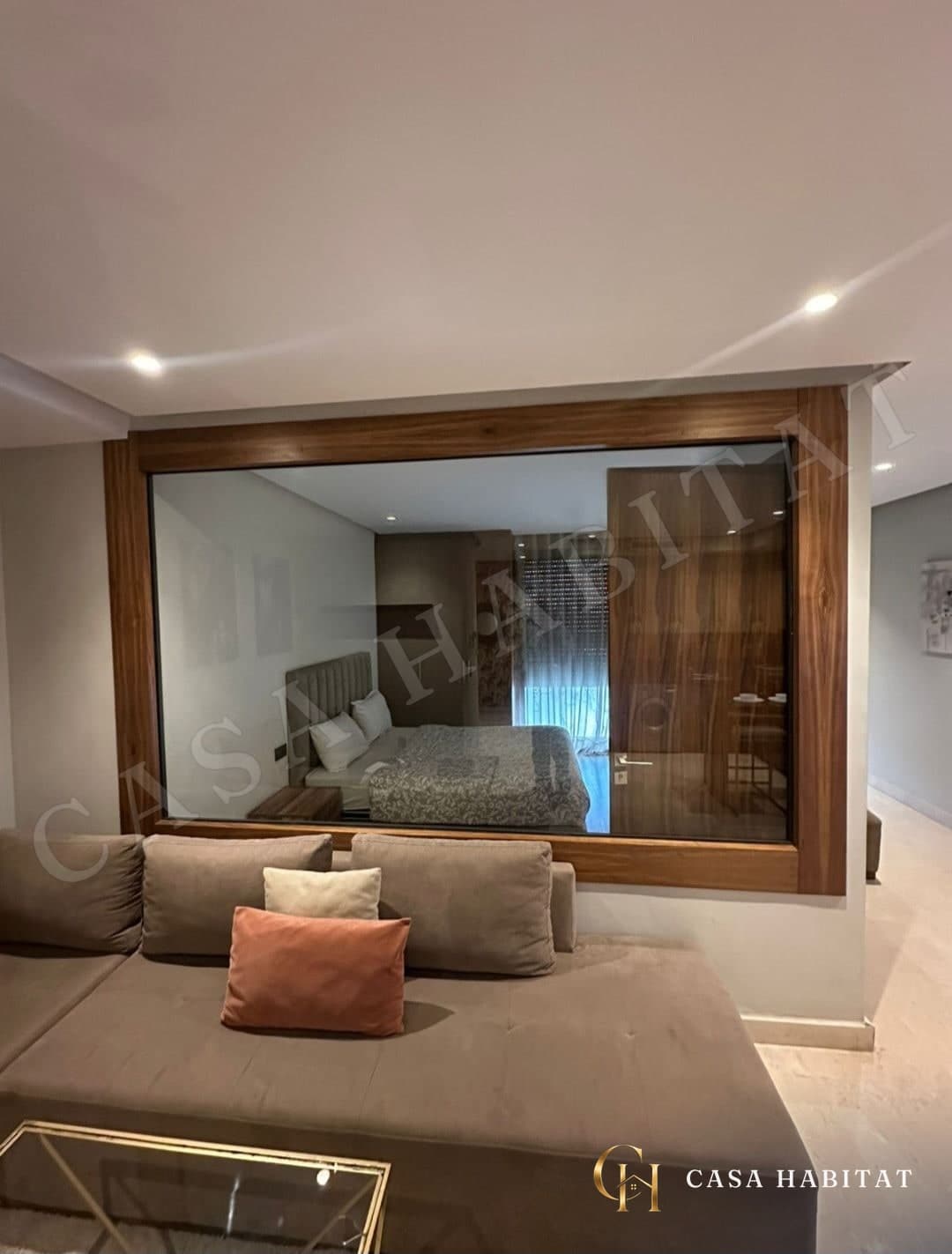 Salon avec canapé beige et grande fenêtre encadrée de bois donnant sur la chambre