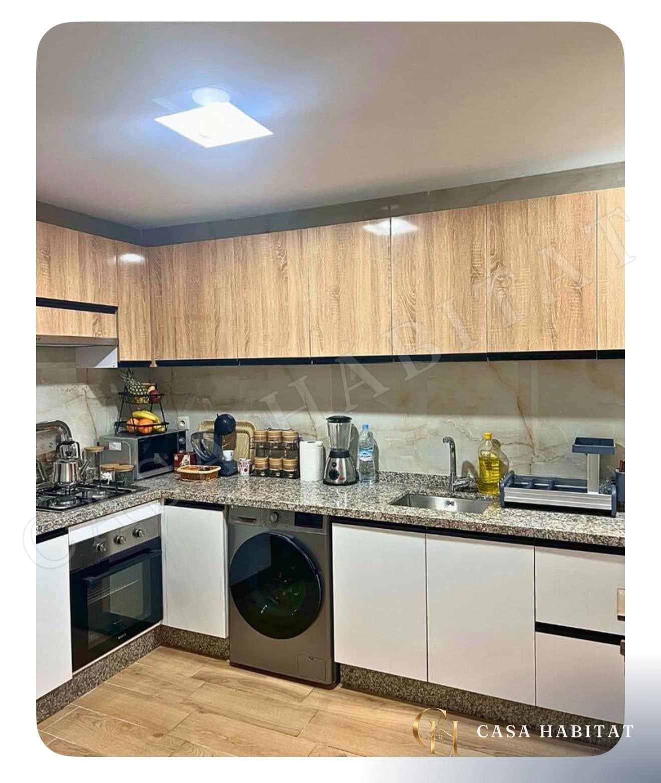 Cuisine avec armoires bois clair, plaque de cuisson, four et lave-linge