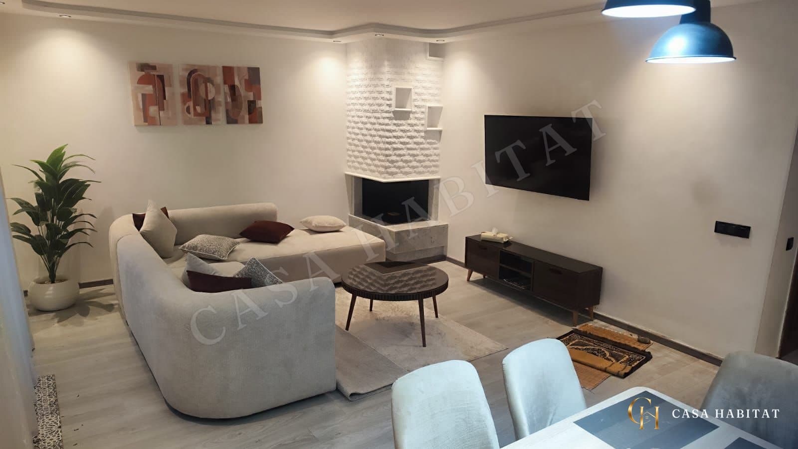 Salon avec canapé d'angle beige, cheminée blanche et téléviseur mural
