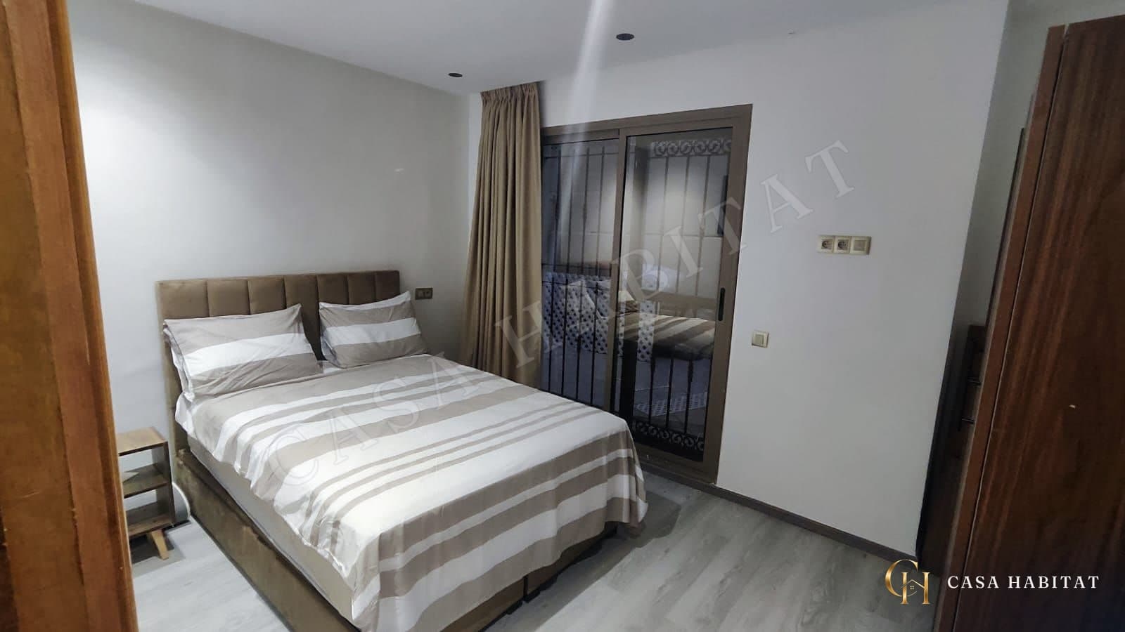 Chambre avec lit double à rayures, rideaux beiges et porte-fenêtre
