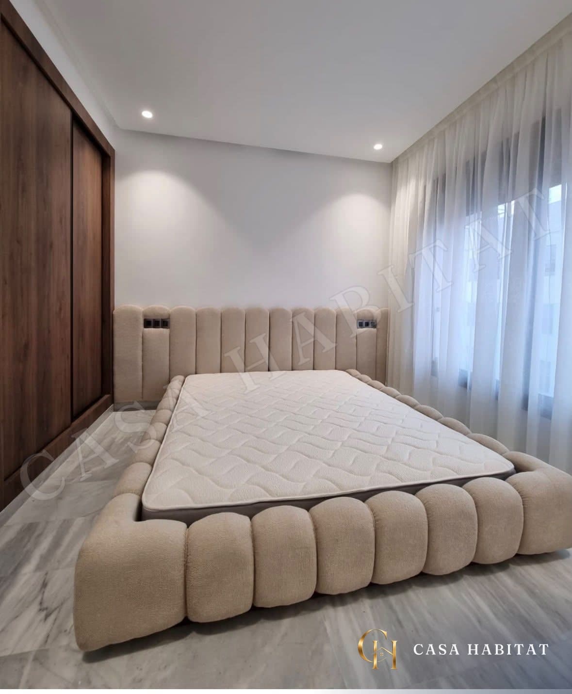 Chambre avec lit capitonné beige, armoire en bois et rideaux clairs