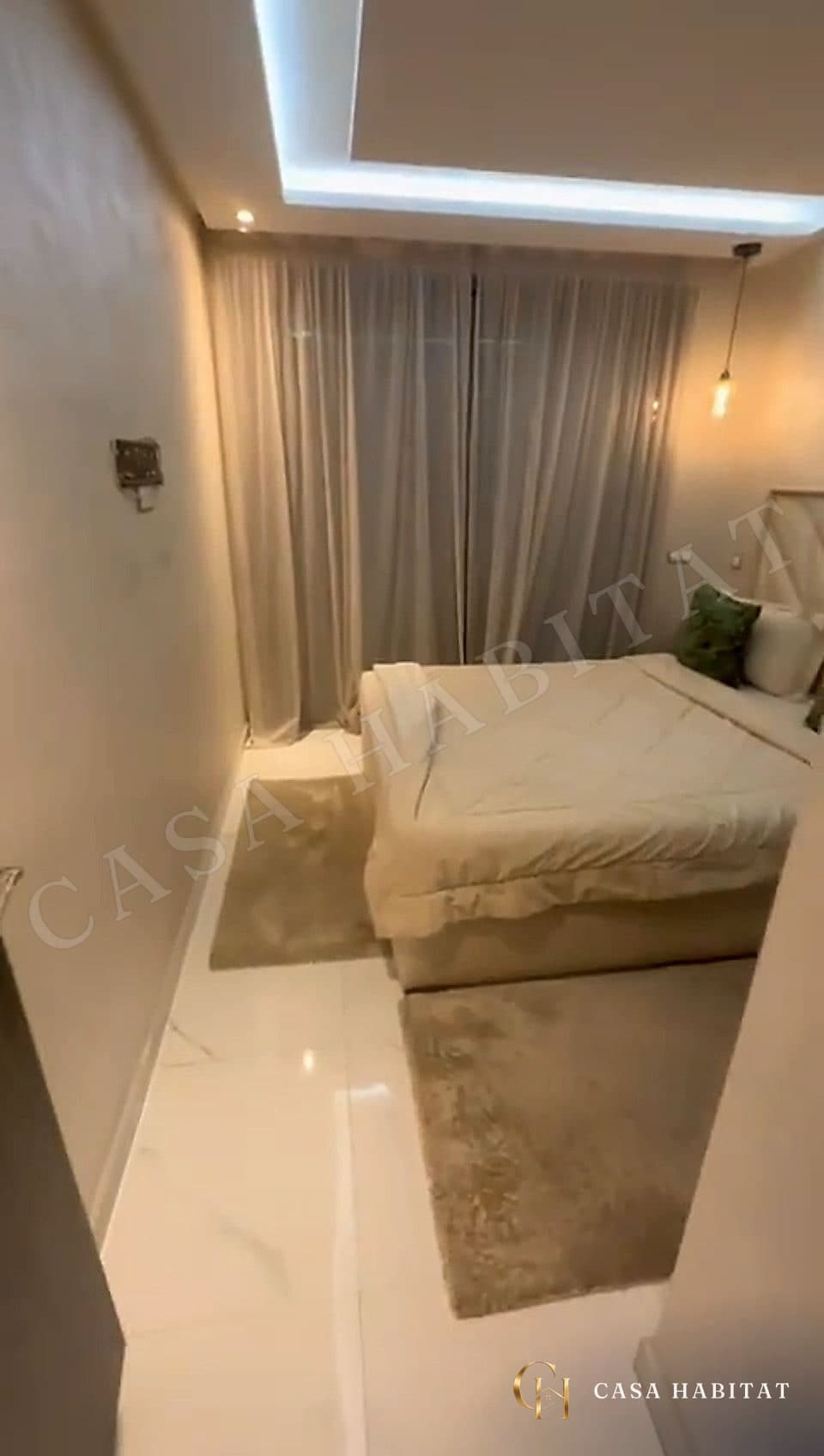 Chambre avec lit beige, petits tapis et lampe suspendue