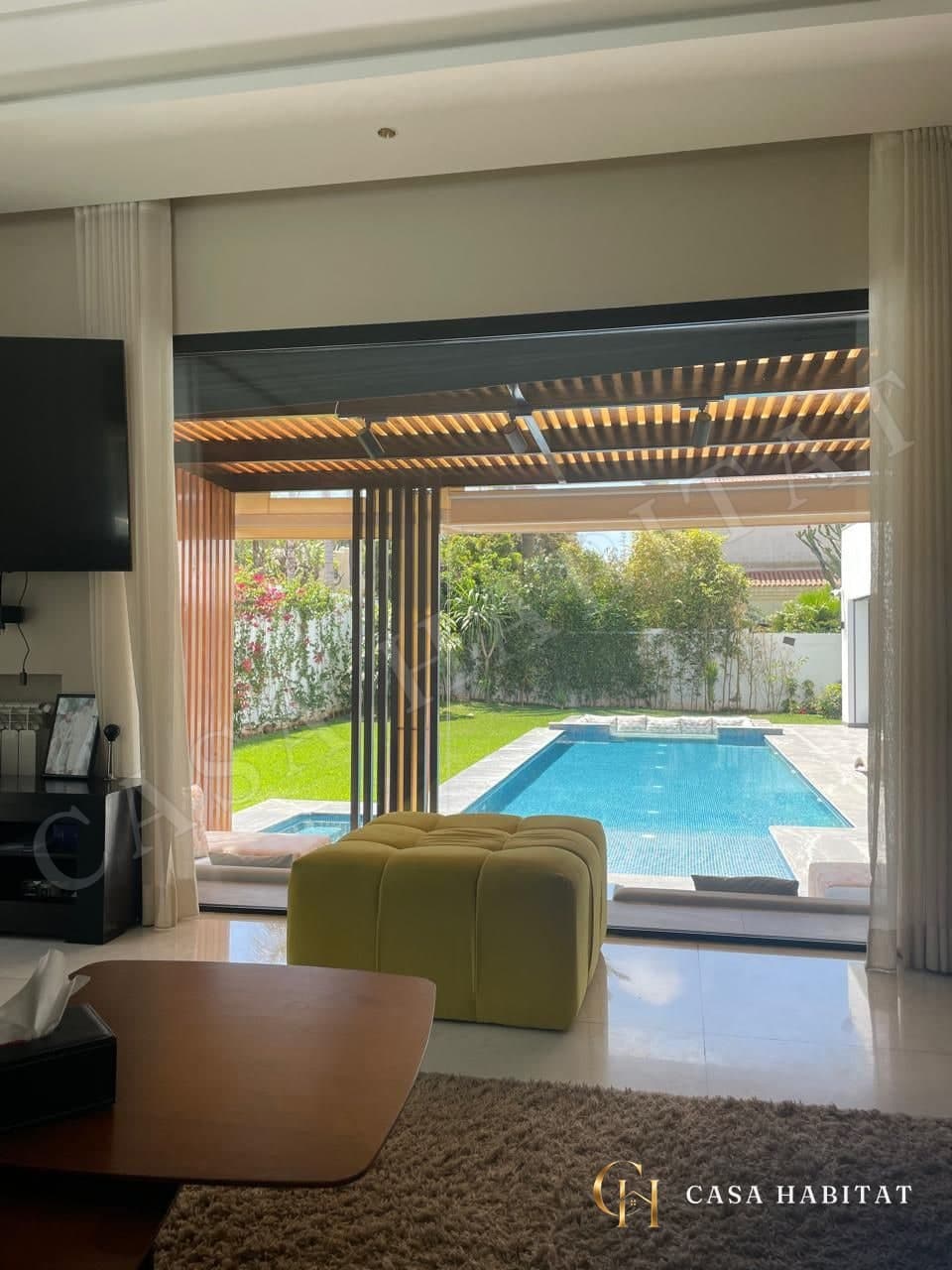 Salon avec pouf jaune donnant sur la piscine et pergola en bois