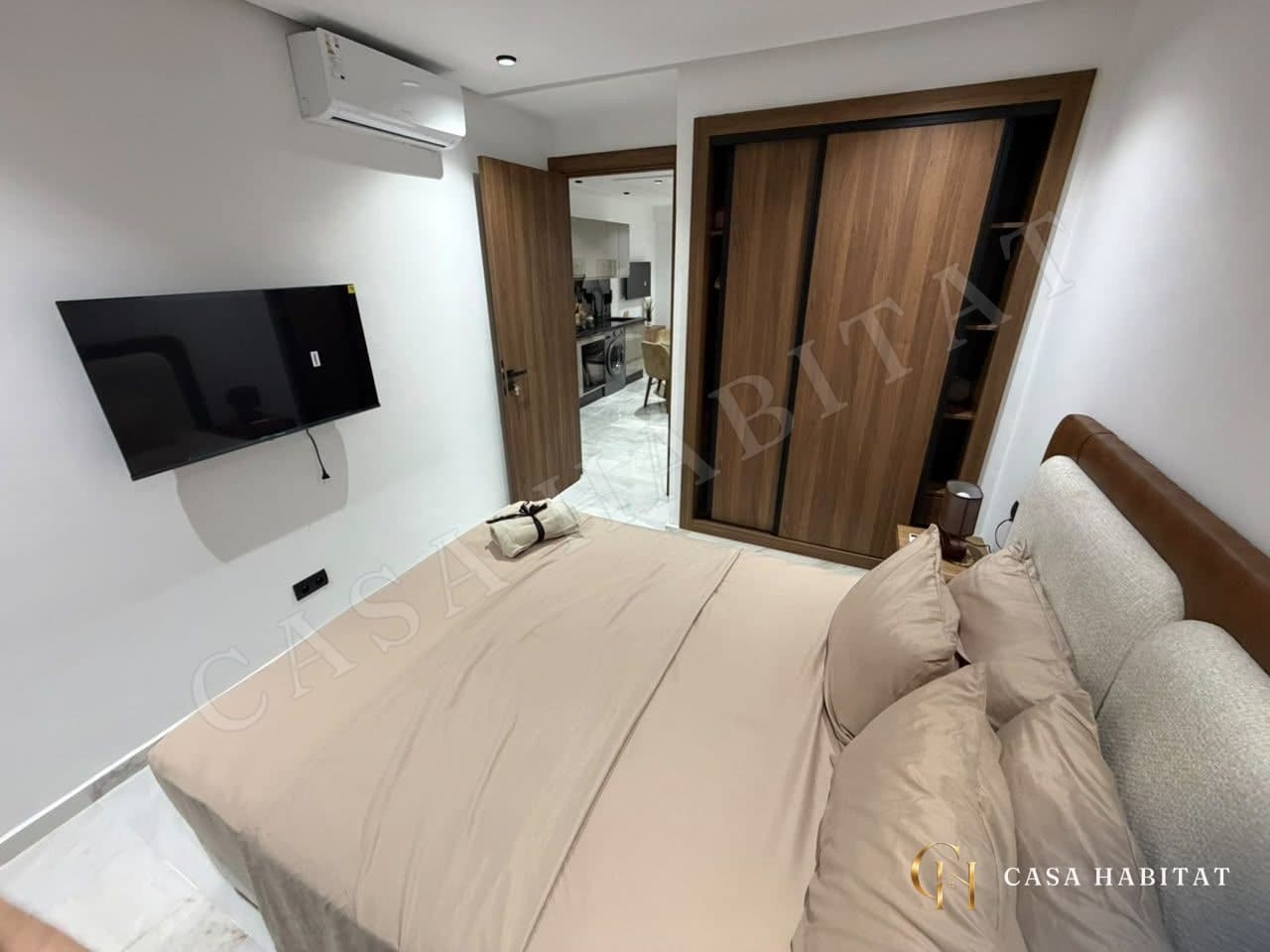 Chambre avec lit beige, climatiseur mural et téléviseur fixé au mur