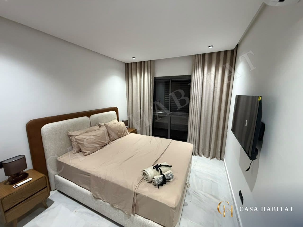 Chambre avec lit à tête capitonnée, rideaux beiges et téléviseur mural