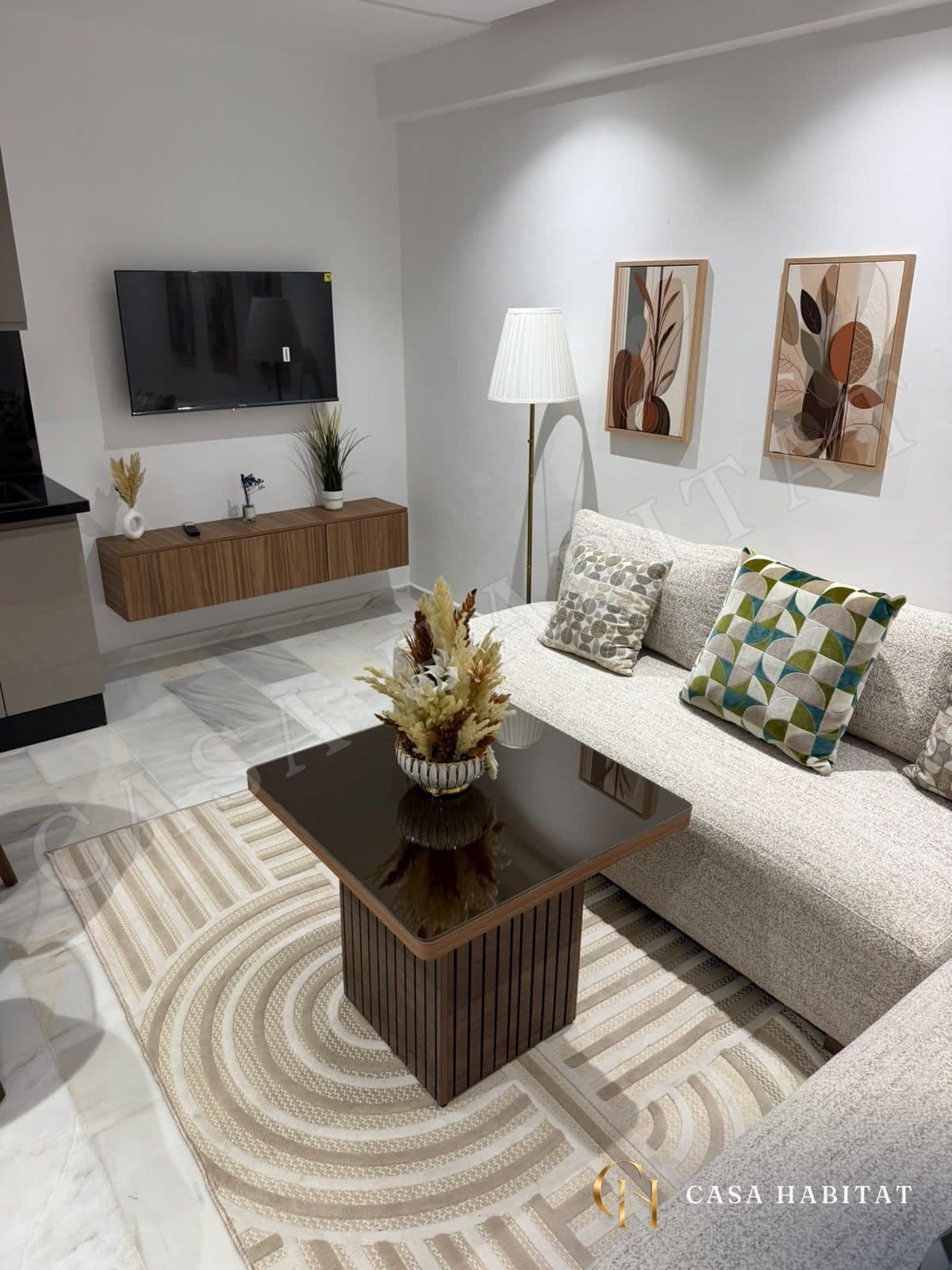 Salon avec canapé beige, téléviseur mural et lampadaire près des tableaux